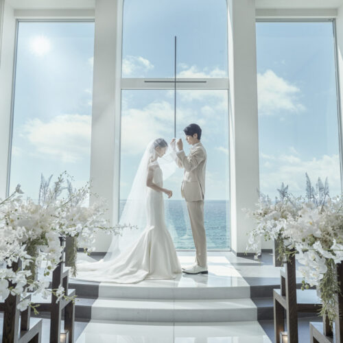 Bride and groom exchange vows at an arched window altar, ocean view beyond, framed by abundant white florals.