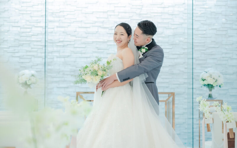 Bride in a white wedding gown holds a bouquet and smiles as the groom wraps his arms around her in a modern venue backdrop.