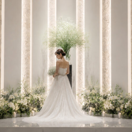 Bride in a strapless white wedding gown standing with her back to the camera in a modern chapel, surrounded by tall stone columns, candles, and white florals.