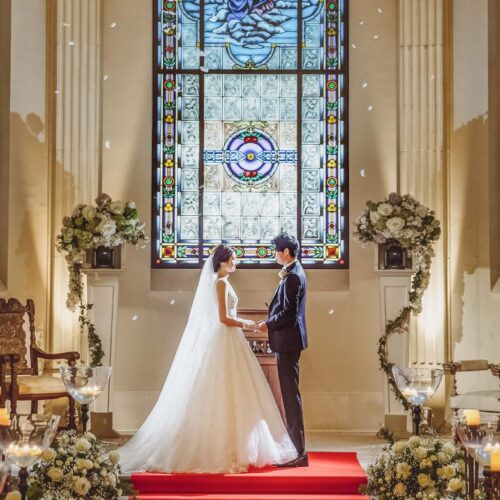 Couple in wedding attire holding hands on a red carpet altar in a church with tall stained glass windows behind them.